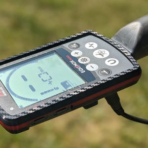 May include: A Minelab Equinox 800 metal detector with a digital display showing readings. The device has a black and red casing with various control buttons. The screen displays a numerical reading and a graphic.