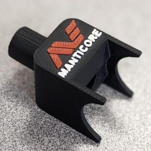 Op de afbeelding: Een zwart 3D-geprint object met het woord "MANTICORE" in witte letters en een rood logo. Het object heeft een cilindrische basis en een rechthoekige bovenkant met gebogen randen. Het logo is een gestileerde "M".