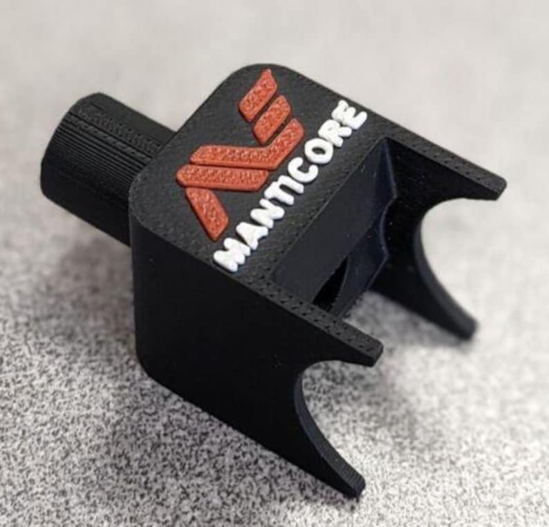 Op de afbeelding: Een zwart 3D-geprint object met het woord "MANTICORE" in witte letters en een rood logo. Het object heeft een cilindrische basis en een rechthoekige bovenkant met gebogen randen. Het logo is een gestileerde "M".