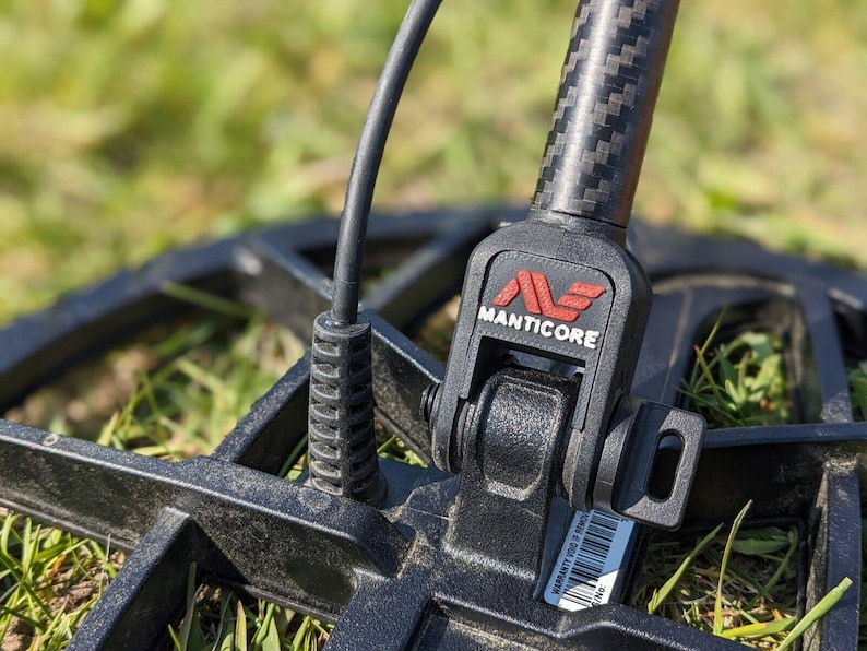 Op de afbeelding: Close-up van een zwarte metaaldetector met de merknaam "MANTICORE" in rood en wit. De detector heeft een koolstofvezel schacht en is gepositioneerd boven gras.