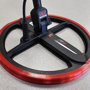 May include: Close-up of a metal detector coil. The coil is black with a red rim. The detector's arm is black, with the brand name "MANTICORE" visible. The model name "M9" is also visible on the coil.