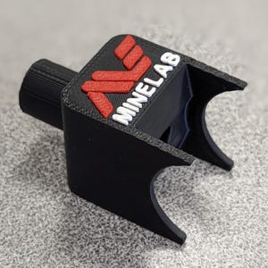 Op de afbeelding: Een zwart 3D-geprint apparaat met het woord "MINELAB" in het wit en een rood logo. Het apparaat heeft een cilindrisch onderdeel en een gebogen, open uiteinde. Het logo staat op een zwarte, rechthoekige basis.