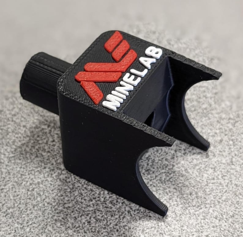 Op de afbeelding: Een zwart 3D-geprint apparaat met het woord "MINELAB" in het wit en een rood logo. Het apparaat heeft een cilindrisch onderdeel en een gebogen, open uiteinde. Het logo staat op een zwarte, rechthoekige basis.