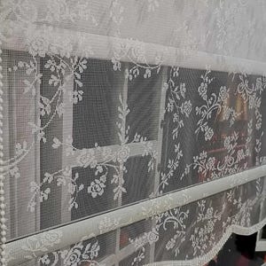 Könnte beinhalten: Weiße Spitzen-Fenstervorhänge mit floralem Muster. Die Vorhänge sind halbtransparent und lassen Licht durch. Das Design umfasst detaillierte Blumen- und Blattmotive, die eine elegante und zarte Ästhetik schaffen. Die Vorhänge sind geschichtet, mit einem gewellten Rand.