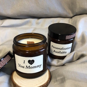 Może przedstawiać: Dwie brązowe świece w szklanych słoikach z czarnymi pokrywkami. Jedna świeca ma etykietę z napisem "I ❤️ You Mommy". Druga świeca ma etykietę z napisem "Personalizacja dostępna". Świece stoją na drewnianej podstawce.