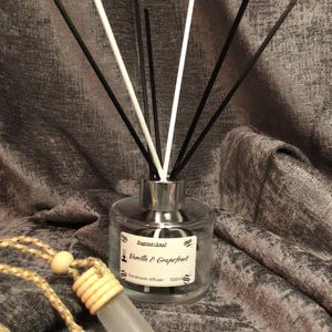 Puede incluir: Un frasco difusor de vidrio transparente con varillas negras y blancas, etiquetado como "Vanilla & Grapefruit". Un pequeño ambientador para coche de vidrio esmerilado con un acento de madera cuelga cerca. El fondo es una tela gris texturizada.