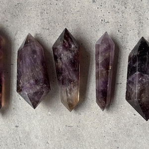 May include: Five double-terminated amethyst crystals arranged in a row. Each crystal features a hexagonal shape with pointed ends, displaying deep purple and translucent violet hues with natural internal inclusions and variations in color intensity.