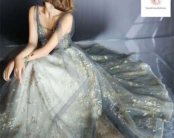 Könnte beinhalten: Ein Abendkleid mit tiefem Ausschnitt und fließendem Rock. Das Kleid hat eine hellfarbene Basis mit aufwendiger goldener Stickerei und ist mit einem transparenten, graublauen Stoff überzogen. Das Kleid ist für besondere Anlässe konzipiert.