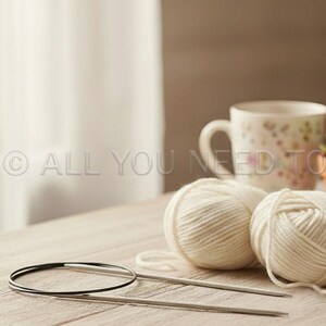 May include: Two balls of cream-colored yarn with knitting needles and a circular needle sit on a wooden surface. A floral-patterned mug, a small potted succulent, and a blue mug are in the background. The image has the text "ALL YOU NEED TO KNIT".