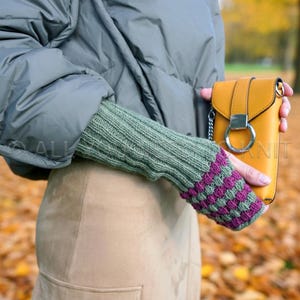 May include: A mustard yellow crossbody bag with a silver ring clasp is held by a person wearing a green knit arm warmer with purple and green striped detailing. The person is wearing a grey puffer jacket and a tan skirt.