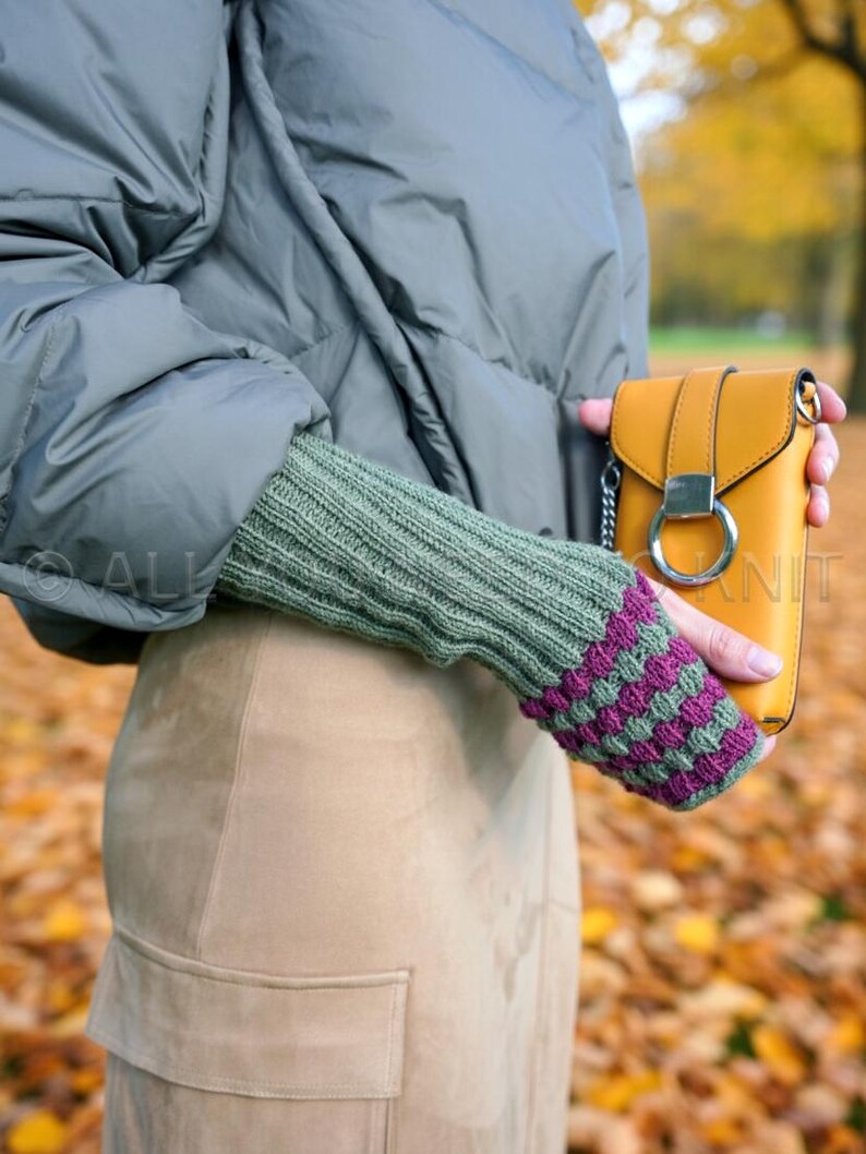 May include: A mustard yellow crossbody bag with a silver ring clasp is held by a person wearing a green knit arm warmer with purple and green striped detailing. The person is wearing a grey puffer jacket and a tan skirt.