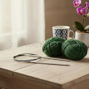 May include: Two balls of dark green yarn and a pair of circular knitting needles rest on a wooden table. A patterned mug and a potted orchid are in the background. The scene is lit by natural light.