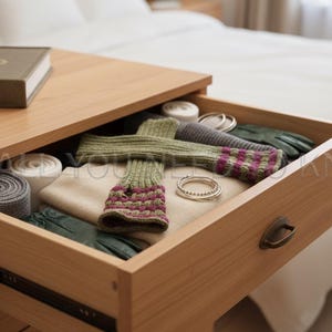 May include: A wooden drawer filled with various items. The drawer contains a pair of green and purple striped fingerless gloves, a beige sweater, green gloves, and silver bracelets. A book rests on the wooden surface above the drawer.