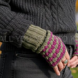 May include: Close-up of a person wearing a dark grey knit sweater and olive green and magenta fingerless gloves. The gloves have a textured, patterned design. The sweater has a cable knit design.