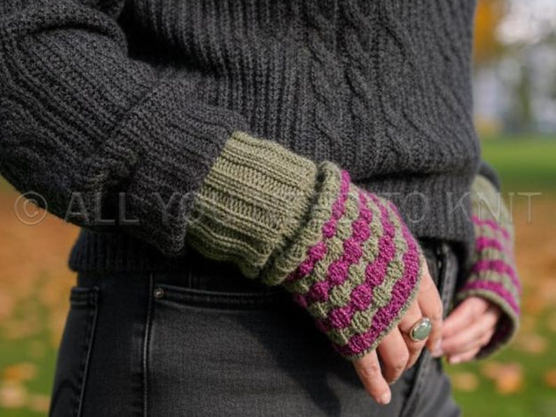May include: Close-up of a person wearing a dark grey knit sweater and olive green and magenta fingerless gloves. The gloves have a textured, patterned design. The sweater has a cable knit design.