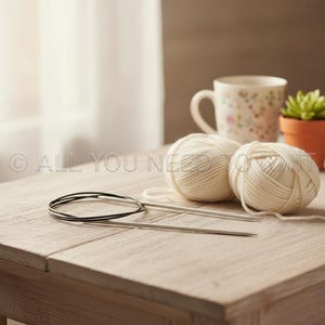 May include: A still-life image featuring knitting supplies on a wooden table. Two balls of cream-colored yarn, a set of circular knitting needles, and a floral-patterned mug are visible. A small potted succulent adds a touch of green.