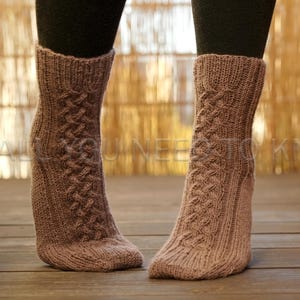 May include: A pair of knitted socks in a muted mauve color. The socks feature a cable knit pattern running down the front and are worn over black leggings. The socks are on a wooden surface.