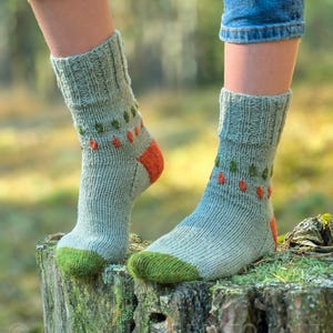 Könnte beinhalten: Ein Paar hell salbeigrüne Strickstrümpfe mit geripptem Bündchen, einer einfarbigen olivgrünen Spitze und einer einfarbigen orangefarbenen Ferse. Der obere Teil der Socken zeigt ein dekoratives Muster aus kleinen, abwechselnden olivgrünen und orangen Stichen.