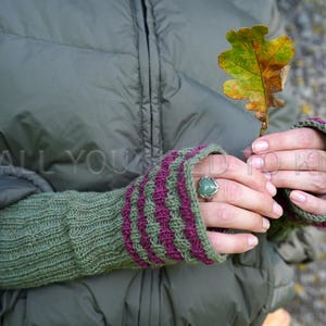 May include: Green and purple knitted fingerless gloves with a textured pattern. The gloves are worn, and a person is holding a colorful leaf. A ring with a green stone is on the person's finger.