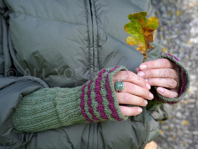 May include: Green and purple knitted fingerless gloves with a textured pattern. The gloves are worn, and a person is holding a colorful leaf. A ring with a green stone is on the person's finger.
