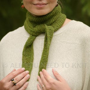 May include: A moss green knitted neck scarf with a textured stitch pattern, tied in a simple knot around the neck. The scarf is worn over a cream-coloured knit jumper. The text ALL YOU NEED TO KNIT is visible across the centre of the image.