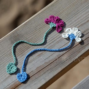 May include: Two crochet flower bookmarks. One features a magenta flower with a light green stem and a small green leaf at the end. The other features a white flower with a blue center, a blue stem, and a small blue leaf at the end of the stem.