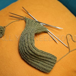 May include: Olive green knitted item in progress, on an orange cushion. The project is partially complete, with knitting needles inserted. A ball of yarn and a heart-shaped yarn detail are also visible. Text on the pillow reads "ALL YOU NEED TO KNIT".