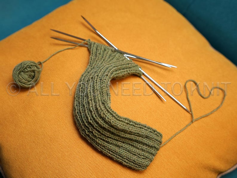 May include: Olive green knitted item in progress, on an orange cushion. The project is partially complete, with knitting needles inserted. A ball of yarn and a heart-shaped yarn detail are also visible. Text on the pillow reads "ALL YOU NEED TO KNIT".