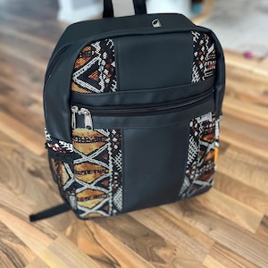 Könnte beinhalten: Schwarzer Rucksack mit oberem Griff und Fronttasche mit Reißverschluss. Der Rucksack hat Stoffpaneele mit orangefarbenen, braunen und weißen geometrischen Mustern. Die Tasche hat einen schwarzen Reißverschluss und ein kleines silberfarbenes Metalldetail.