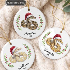 May include: Three round white ceramic ornaments featuring illustrations of snakes wearing red and white Santa hats, framed by green holly leaves and red berries. Each ornament is personalized with a name and the year 2026 in black script.