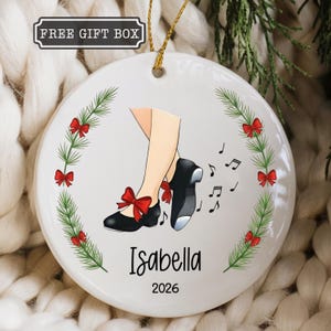 May include: A round white ceramic ornament featuring a graphic of legs in black tap shoes with red bows. The design includes a wreath of green pine branches with red bows, the name Isabella, the year 2026, and musical notes. A gold string is attached.