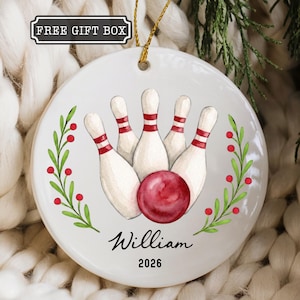 May include: A round white ceramic ornament featuring a watercolor illustration of five bowling pins and a red bowling ball. The design includes green leafy branches with red berries on both sides, the name William, and the year 2026 in black text.
