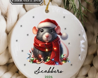 May include: A round white ceramic ornament featuring a gray rat wearing a red Santa hat and a red scarf, tangled in colorful Christmas lights. The ornament includes the name Scabbers and the year 2026 printed in black and green text.