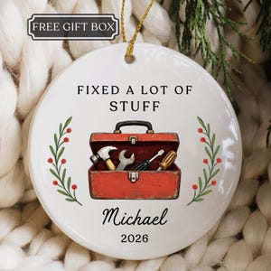 May include: A round white ceramic ornament featuring a red toolbox illustration with a hammer, wrench, and screwdriver. Text reads "FIXED A LOT OF STUFF", "Michael", and "2026". Green sprigs with red berries flank the toolbox, and a gold string is attached.