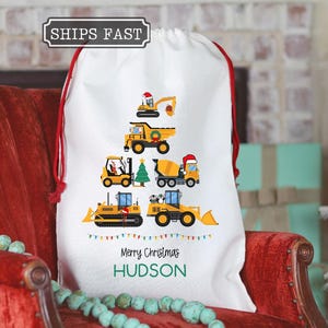 May include: White Christmas gift bag with a red drawstring, featuring a design of yellow construction vehicles arranged in a Christmas tree shape. The bag is personalized with the name "HUDSON" and the text "Merry Christmas".