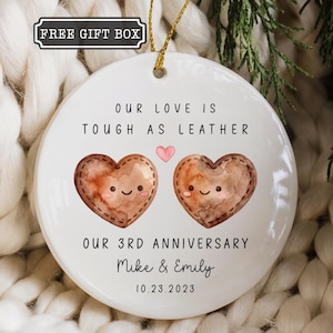 May include: A round white ceramic ornament featuring the text "OUR LOVE IS TOUGH AS LEATHER" and "OUR 3RD ANNIVERSARY" with the names "Mike & Emily" and the date "10.23.2023". Two brown watercolor hearts with faces are centered with a small pink heart.