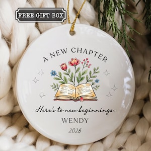 May include: A round white ceramic ornament featuring a gold hanging string. The design includes an open book with colorful flowers emerging from the pages, surrounded by star icons. Text reads: A NEW CHAPTER, Here's to new beginnings, WENDY, 2026.