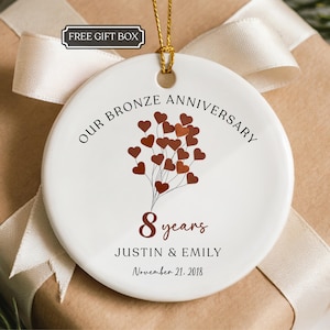 May include: A round white ceramic ornament featuring a cluster of bronze-colored heart balloons, the text "OUR BRONZE ANNIVERSARY", the number "8 years", the names "JUSTIN & EMILY", and the date "November 21, 2018" in black and bronze lettering.