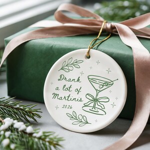 May include: A round white ceramic ornament featuring a green line drawing of a martini glass with olives and a bow. The text reads Drank a lot of Martinis 2026. It includes green olive branch illustrations and small star accents, suspended by a gold cord.