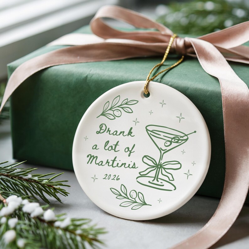 May include: A round white ceramic ornament featuring a green line drawing of a martini glass with olives and a bow. The text reads Drank a lot of Martinis 2026. It includes green olive branch illustrations and small star accents, suspended by a gold cord.