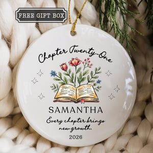 May include: A round white ceramic ornament featuring a gold hanging string. The design includes an open book with colorful flowers emerging from the pages, the text "Chapter Twenty-One", "SAMANTHA", "Every chapter brings new growth.", and "2026".