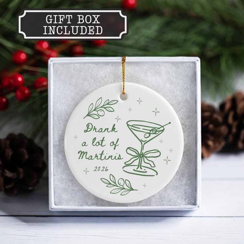 May include: A round white ceramic ornament featuring a green line drawing of a martini glass with olives, a bow, and the text "Drank a lot of Martinis 2026". The ornament is suspended by a gold string and is presented inside a white gift box.