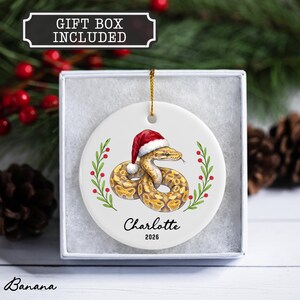 May include: A round white ceramic ornament featuring a coiled snake wearing a red and white Santa hat, flanked by green holly branches with red berries. The text "Charlotte" and "2026" is printed below the snake. A gold string is attached.
