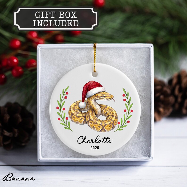 May include: A round white ceramic ornament featuring a coiled snake wearing a red and white Santa hat, flanked by green holly branches with red berries. The text "Charlotte" and "2026" is printed below the snake. A gold string is attached.