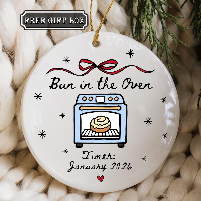 K&ouml;nnte beinhalten: Runder wei&szlig;er Keramikschmuck mit goldenem Faden. Der Schmuck zeigt den Text "Bun in the Oven" &uuml;ber einem Cartoon-Ofen mit einem Backwaren darin. Der Text "Timer: January 2026" steht darunter, mit einem roten Herz. Der Text "FREE GIFT BOX" steht in einer schwarzen Box.