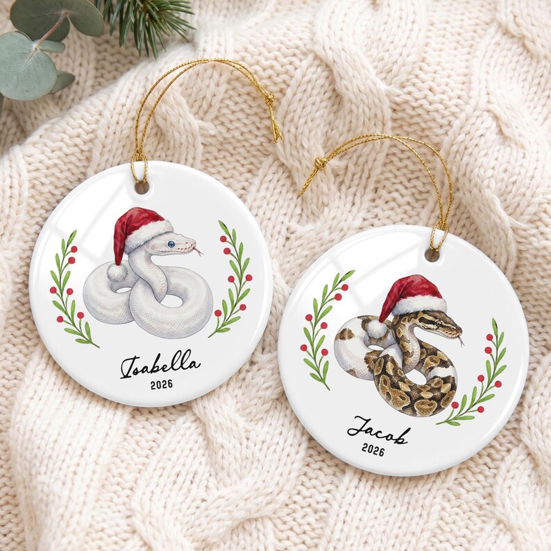 May include: Two round white ceramic ornaments featuring illustrations of snakes wearing red and white Santa hats. One snake is white, the other is brown and tan. Both are framed by green holly branches with red berries. Text reads Isabella 2026 and Jacob 2026.