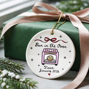 K&ouml;nnte beinhalten: Wei&szlig;er Keramik-Weihnachtsschmuck mit dem Text "Bun in the Oven" und einer rosa Ofen-Grafik. Der Schmuck enth&auml;lt den Text "Timer: January 2026" und ist mit kleinen Sternen und einer roten Schleife verziert. Ein goldener Faden ist zum Aufh&auml;ngen befestigt.