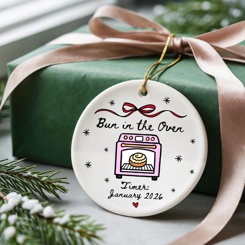 K&ouml;nnte beinhalten: Wei&szlig;er Keramik-Weihnachtsschmuck mit dem Text "Bun in the Oven" und einer rosa Ofen-Grafik. Der Schmuck enth&auml;lt den Text "Timer: January 2026" und ist mit kleinen Sternen und einer roten Schleife verziert. Ein goldener Faden ist zum Aufh&auml;ngen befestigt.