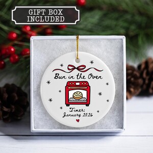 K&ouml;nnte beinhalten: Wei&szlig;es Keramikornament mit einer roten Ofengrafik und dem Text "Bun in the Oven". Enth&auml;lt den Text "Timer: January 2026". Das Ornament befindet sich in einer Geschenkbox und hat eine goldene Schnur zum Aufh&auml;ngen.