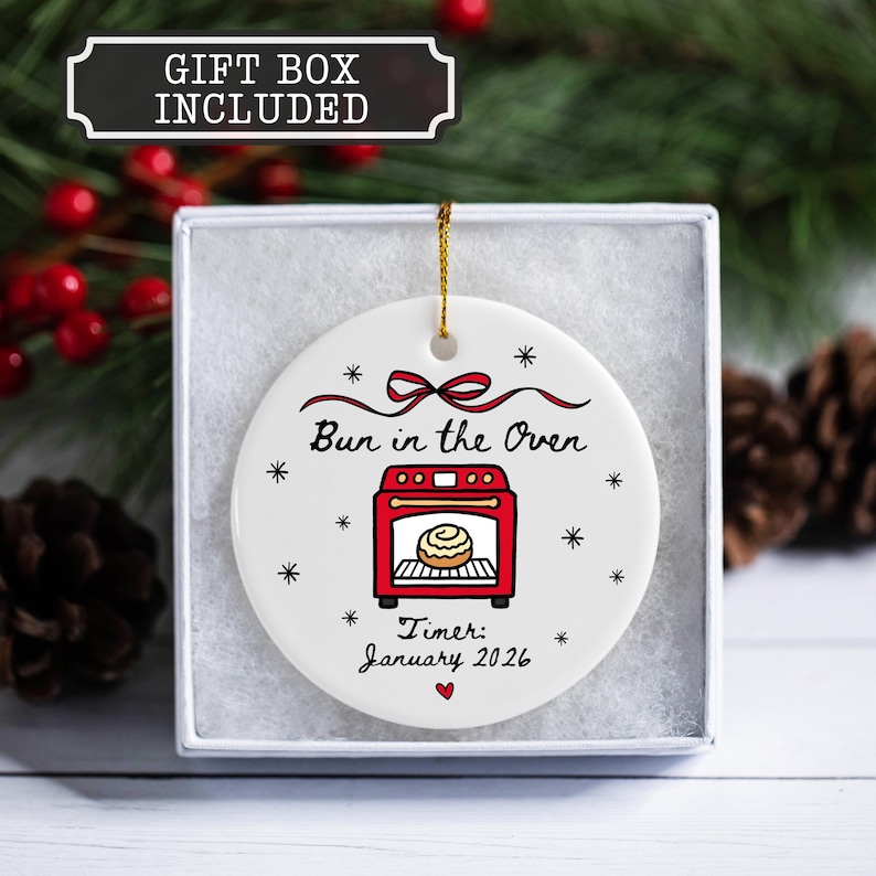 K&ouml;nnte beinhalten: Wei&szlig;es Keramikornament mit einer roten Ofengrafik und dem Text "Bun in the Oven". Enth&auml;lt den Text "Timer: January 2026". Das Ornament befindet sich in einer Geschenkbox und hat eine goldene Schnur zum Aufh&auml;ngen.